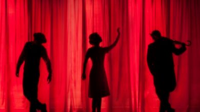 Auf dem Bild ist eine Bühne mit rotem Vorhang zu sehen mit drei Personen im Schatten.