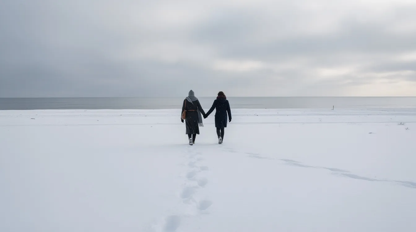 Zwei Personen, Hand in Hand, laufen über eine menschenleere, verschneite Strandlandschaft, während im Hintergrund ein grauer Winterhimmel und ein ruhiges Meer zu sehen sind. Diese Szene erinnert an die melancholische Atmosphäre von "Eternal Sunshine of the Spotless Mind", wo Erinnerungen und Beziehungen im Mittelpunkt stehen.